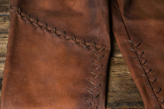 Close view of brown leather trouser panels with hand-laced seams on a wooden surface