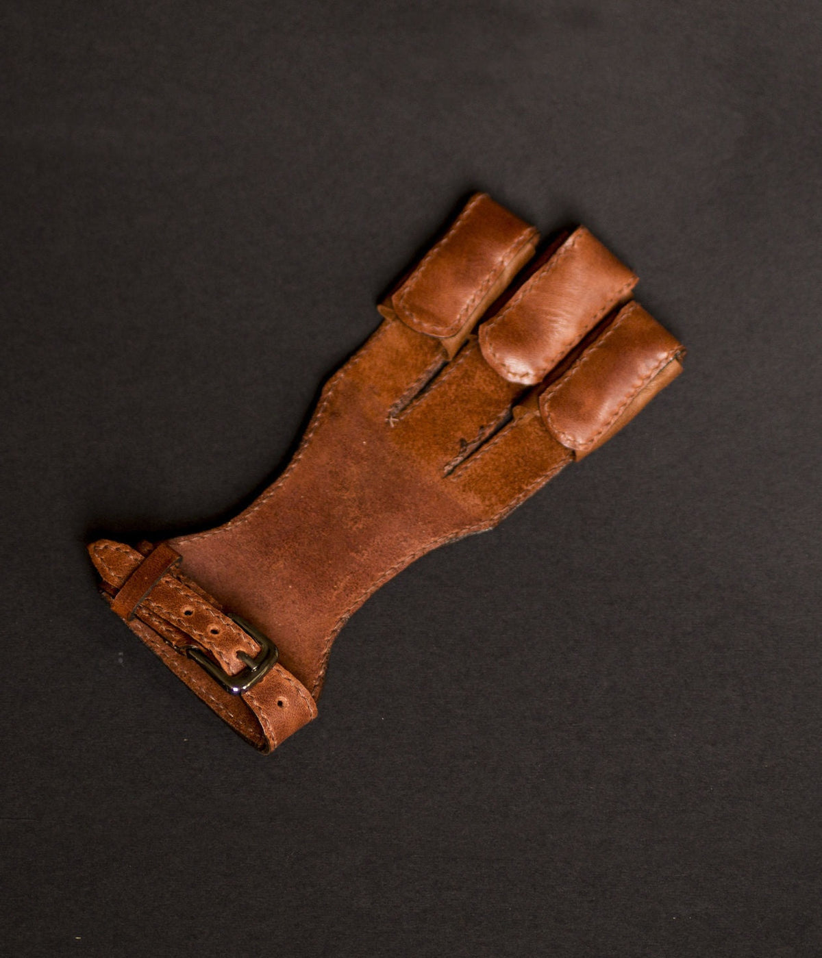 Brown right-hand leather archery glove with three finger guards on a dark background