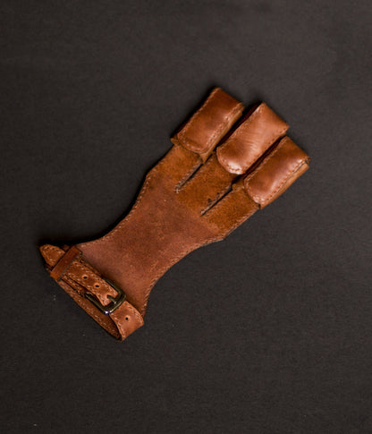 Brown right-hand leather archery glove with three finger guards on a dark background
